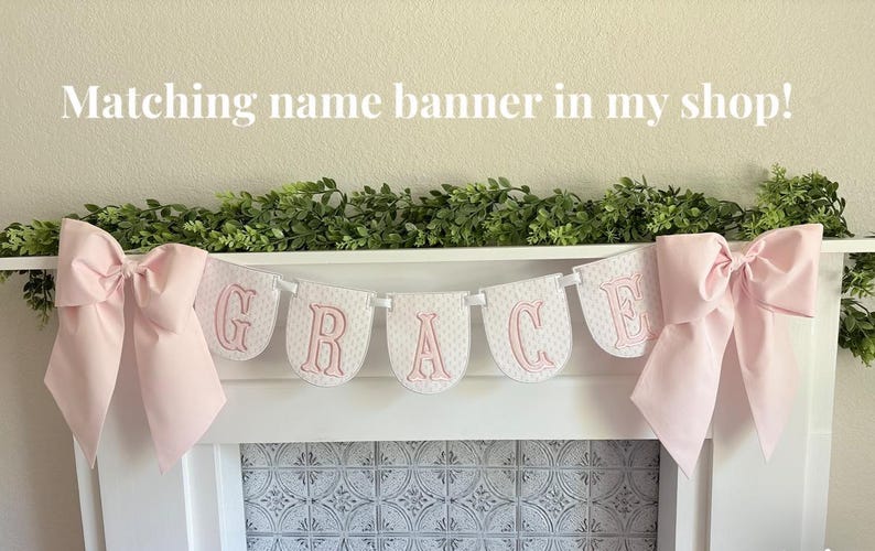 May include: A white banner spelling "GRACE" in pink letters, with a pink bow on each end. The banner is strung across a white shelf with greenery above. The text "Matching name banner in my shop!" is at the top.