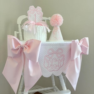 May include: A white high chair decorated for a first birthday. It features a pink bow on each side, a pink and white patterned fabric cover, and a cake with a bow-shaped topper. A pink pom-pom party hat sits next to the cake.