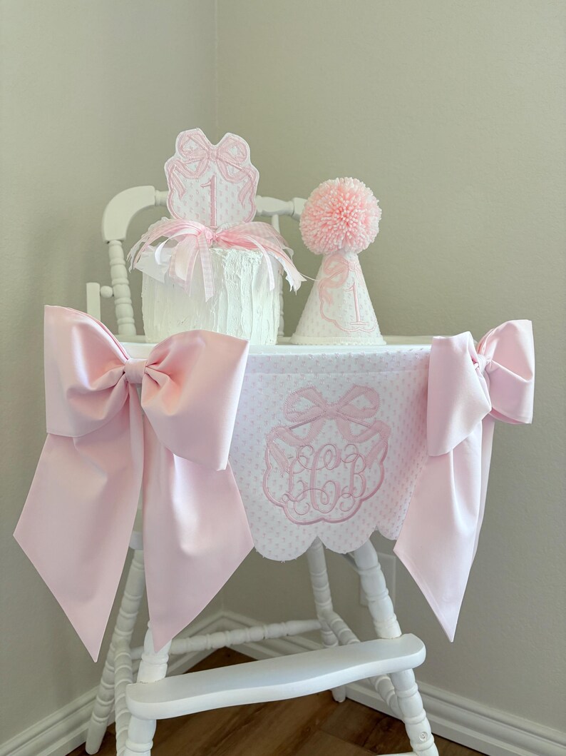 May include: A white high chair decorated for a first birthday. It features a pink bow on each side, a pink and white patterned fabric cover, and a cake with a bow-shaped topper. A pink pom-pom party hat sits next to the cake.