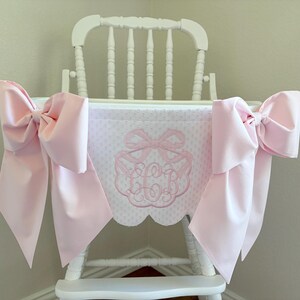 May include: A white wooden high chair with a pink bow and a pink and white patterned fabric. The fabric has a pink embroidered monogram. Large pink bows are attached to the sides of the high chair.