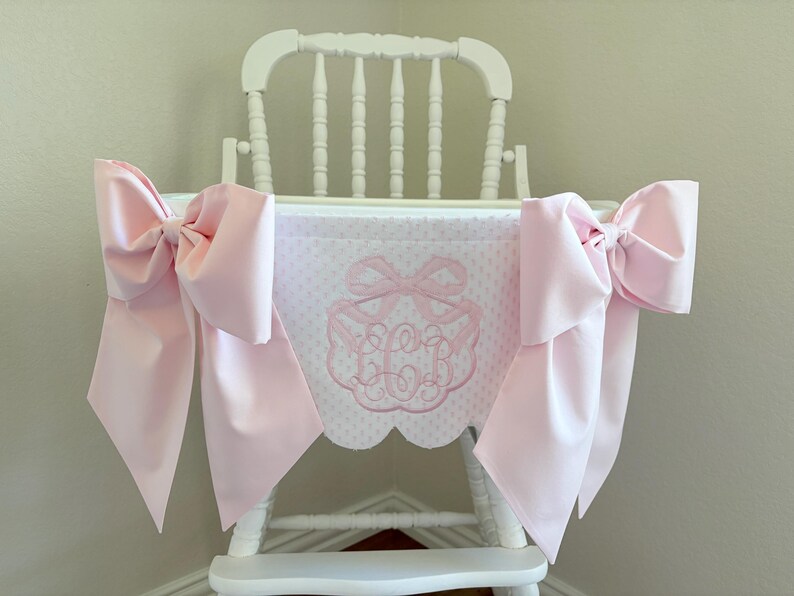 May include: A white wooden high chair with a pink bow and a pink and white patterned fabric. The fabric has a pink embroidered monogram. Large pink bows are attached to the sides of the high chair.