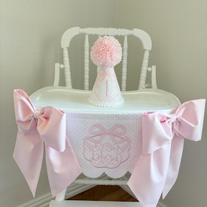 May include: A white high chair decorated with a pink bow-adorned banner and matching bows. A pink party hat with a pom-pom and the number 1 sits on the tray. The banner features a monogram and polka dots.