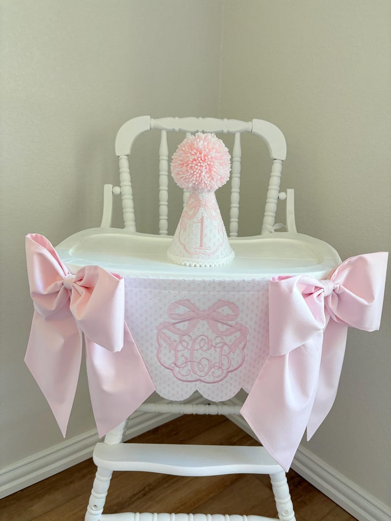 May include: A white high chair decorated with a pink bow-adorned banner and matching bows. A pink party hat with a pom-pom and the number 1 sits on the tray. The banner features a monogram and polka dots.