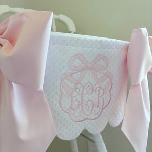 May include: A white high chair cover with a pink bow and monogram embroidery. The cover has a scalloped edge and is adorned with large pink bows on each side. The design features a repeating pattern of small pink dots.