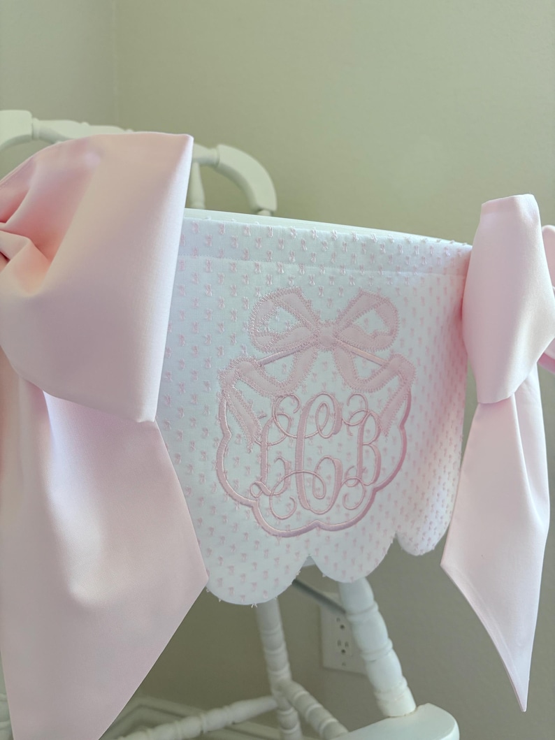 May include: A white high chair cover with a pink bow and monogram embroidery. The cover has a scalloped edge and is adorned with large pink bows on each side. The design features a repeating pattern of small pink dots.