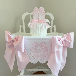 May include: A white high chair decorated with pink bows and a matching table skirt. The skirt features a scalloped edge and an embroidered monogram. A small cake with a bow topper sits on the chair.