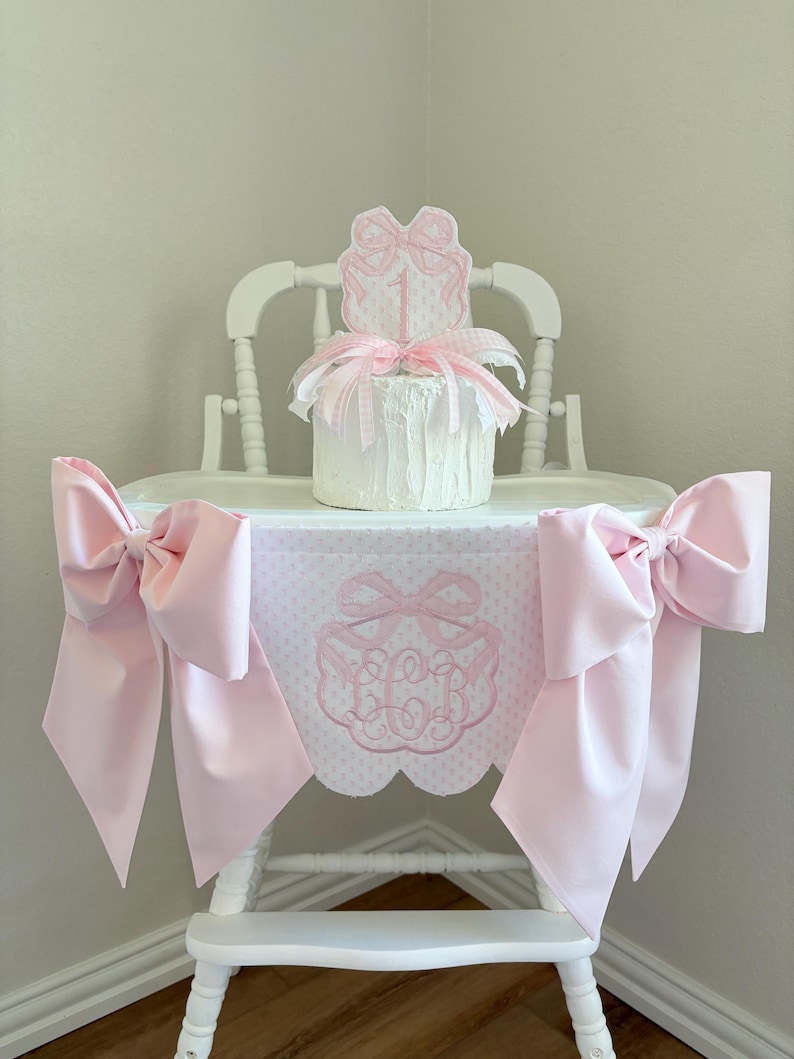 May include: A white high chair decorated with pink bows and a matching table skirt. The skirt features a scalloped edge and an embroidered monogram. A small cake with a bow topper sits on the chair.