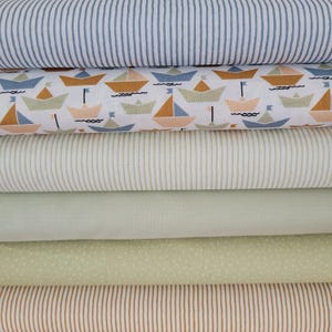 Könnte beinhalten: Ein Stapel verschiedener Stoffmuster. Der obere Stoff ist blau-weiß gestreift. Darunter befindet sich ein weißer Stoff mit einem Muster aus Segelbooten in Orange, Blau und Grün. Weitere Stoffe sind Streifen, einfarbiges Grün und ein gepunktetes Muster.