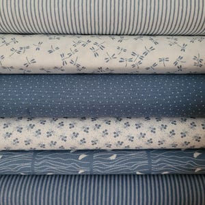 Könnte beinhalten: Ein Stapel aus sechs gefalteten Stoffballen mit blau-weißen Mustern. Die Designs umfassen vertikale Streifen, verstreute blaue Libellen auf weißem Grund, kleine weiße Punkte auf Blau, winzige blaue Blumenmotive sowie wellenförmige Linien mit Vögeln.