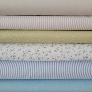 Könnte beinhalten: Ein Stapel Stoffrollen in verschiedenen Mustern und Farben. Die Stoffe umfassen unifarbenes Beige, beige und weiße Streifen, hellgrün mit weißen Punkten, weiß mit blauem Blumendruck, blau und weiße Streifen und unifarbenes Hellblau.