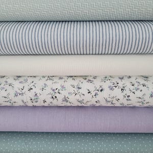 Könnte beinhalten: Ein Stapel Stoffrollen in verschiedenen Mustern und Farben. Obenauf ein hellgrünes Zickzackmuster, gefolgt von blau-weißen Streifen, einfarbigem Weiß, floralem Druck, einfarbigem Lavendel und einem hellgrünen Tupfenmuster.