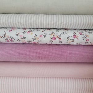 Könnte beinhalten: Ein Stapel Stoffmuster in verschiedenen Farben und Mustern. Die Stoffe umfassen unifarbenes Beige, beige und weiße Streifen, Weiß mit rosa Blumenmuster, unifarbenes Rosa, unifarbenes helles Rosa und hellrosa und weiße Streifen. Die Stoffe sind ordentlich gefaltet.