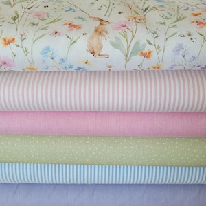 Könnte beinhalten: Ein Stapel Stoffrollen in verschiedenen Mustern und Farben. Die oberste Schicht zeigt einen Blumendruck mit einer Kaninchenillustration. Weitere Schichten umfassen rosa Streifen, einfarbiges Rosa, grüne Punkte, blaue Streifen und einfarbiges Lavendel.