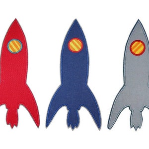 Könnte beinhalten: Drei Filz-Raketen-Patches in Rot, Blau und Grau. Jede Rakete hat einen gelben und orangen Kreis mit roten Streifen in der Mitte.
