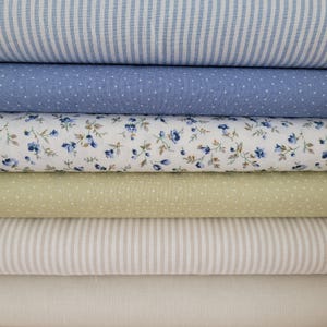 Könnte beinhalten: Ein Stapel Stoffballen in verschiedenen Mustern und Farben. Die Stoffe umfassen blau-weiße Streifen, blau mit weißen Punkten, weiß mit blauem Blumenmuster, hellgrün mit weißen Punkten, beige Streifen und einfarbiges Off-White.