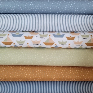 Könnte beinhalten: Ein Stapel Stoffrollen mit verschiedenen nautischen Mustern. Die Stoffe umfassen Blau mit weißen Punkten, blau-weiße Streifen, einen Segelboot-Druck, hellgrün mit weißen Punkten und Senfgelb mit weißen Punkten.