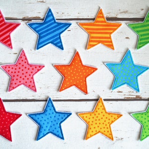 Könnte beinhalten: Zehn Filz-Stern-Patches in verschiedenen Farben und Mustern, darunter Rot, Rosa, Orange, Gelb, Blau, Grün und Weiß. Die Sterne haben einen weißen Umriss und sind in drei Reihen auf einer weißen Holzoberfläche angeordnet.