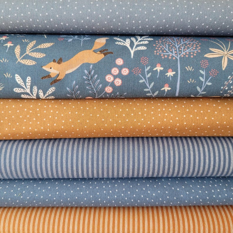 Op de afbeelding: Een stapel van vijf gevouwen stoffen. De bovenste stof is blauw met witte stippen. De tweede is blauw met een vossen- en bloemenprint. De derde is mosterdgeel met witte stippen. De vierde is blauw met witte strepen. De vijfde is blauw met witte stippen. De onderste is mosterdgeel met witte strepen.