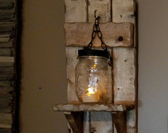 Può includere: Un ripiano a muro in legno rustico con un barattolo di vetro appeso a una catena. Il barattolo contiene una candela accesa. Il ripiano è realizzato in legno riciclato e ha una finitura usurata.