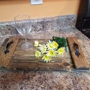 May include: A rustic wooden serving tray with two black metal handles. The tray is stained a dark brown color and has a natural wood grain. A bouquet of yellow silk flowers is placed on the tray, along with a clear glass.