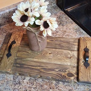 May include: A rustic wooden serving tray with two black metal handles. The tray is made of reclaimed wood and has a natural, distressed finish. A mason jar filled with dried flowers sits on the tray.