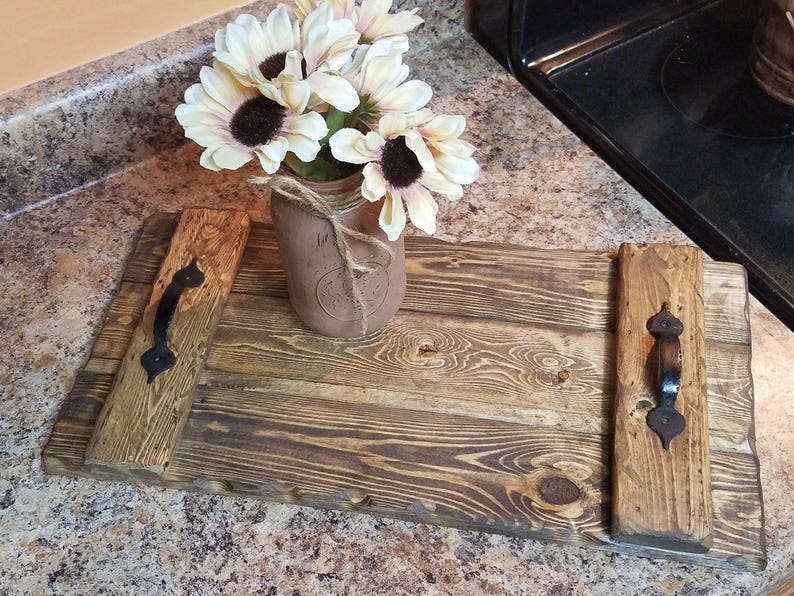 May include: A rustic wooden serving tray with two black metal handles. The tray is made of reclaimed wood and has a natural, distressed finish. A mason jar filled with dried flowers sits on the tray.
