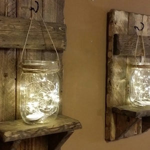May include: Two rustic wooden wall shelves with a glass jar hanging from a rope and a shelf below. Each jar contains a string of fairy lights. The shelves are made of reclaimed wood and have a distressed finish.