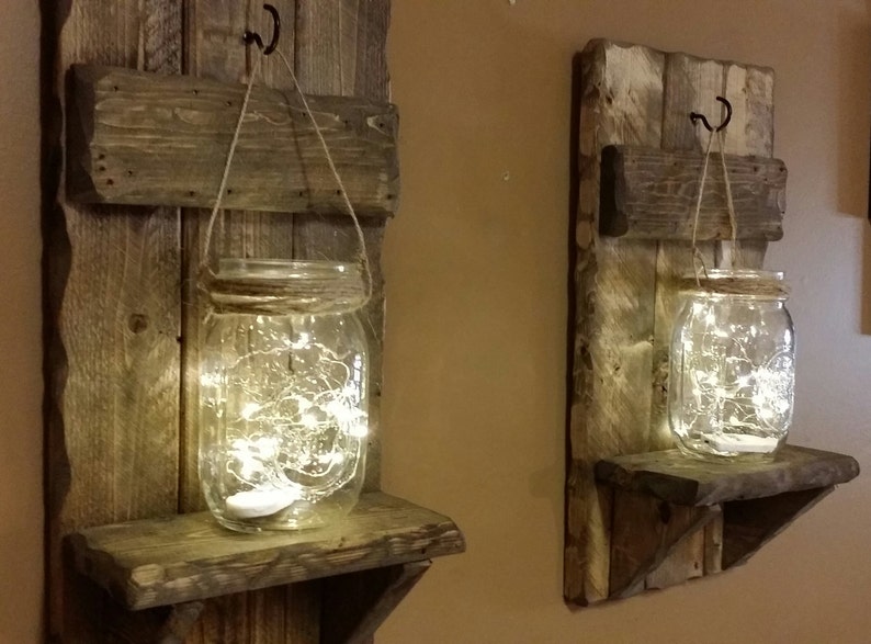 May include: Two rustic wooden wall shelves with a glass jar hanging from a rope and a shelf below. Each jar contains a string of fairy lights. The shelves are made of reclaimed wood and have a distressed finish.