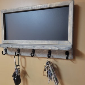 May include: A rustic wooden key holder with a chalkboard panel and five hooks. The hooks are black metal and have keys hanging from them. The wood is painted a light gray with a distressed finish.