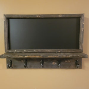 May include: A dark brown wooden shelf with a chalkboard above it. The shelf has four black metal hooks and a rustic finish.