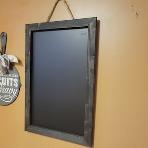 May include: A rectangular chalkboard with a dark gray wooden frame hangs on a wall. The chalkboard is suspended by a natural rope. To the left, a decorative sign in the shape of a skillet reads "We go together like biscuits and gravy."