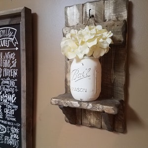May include: A rustic wooden wall shelf with a white painted mason jar filled with faux white flowers. The shelf has a small hook on the top and is stained in a dark brown color. The mason jar has the word "Ball" printed on it.