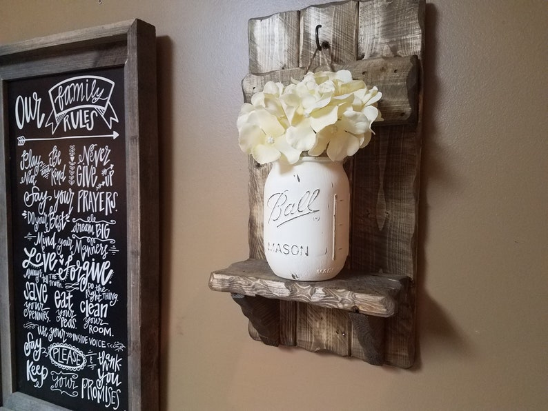 May include: A rustic wooden wall shelf with a white painted mason jar filled with faux white flowers. The shelf has a small hook on the top and is stained in a dark brown color. The mason jar has the word "Ball" printed on it.