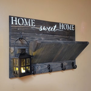 May include: A rustic, dark-stained wooden wall shelf with the words "HOME sweet HOME" in white script. It features a small shelf, four black hooks, and a black lantern with a lit candle, perfect for entryway decor.