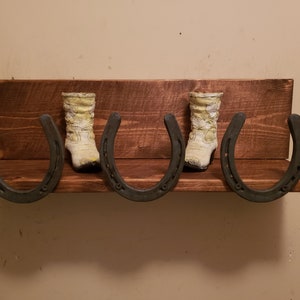 May include: A wooden shelf with three horseshoe hooks, each holding a pair of white cowboy boots. The shelf is stained a dark brown color.