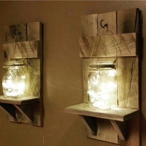 May include: Two rustic wooden wall shelves with a mason jar on each shelf. The jars are filled with fairy lights and hung from a hook on the shelf.