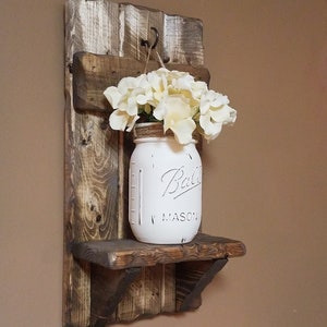 May include: A rustic wooden wall shelf with a white painted mason jar filled with white flowers. The shelf has a small hook on the top and a small shelf on the bottom. The jar has the text "Ball Mason" on it.