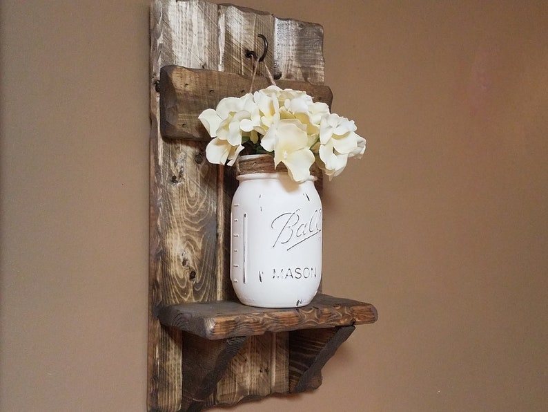 May include: A rustic wooden wall shelf with a white painted mason jar filled with white flowers. The shelf has a small hook on the top and a small shelf on the bottom. The jar has the text "Ball Mason" on it.
