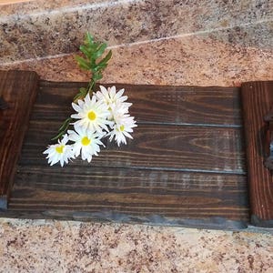 May include: A wooden serving tray with two black metal handles. The tray has a dark brown stain and is made of reclaimed wood. A bouquet of white daisies is on the tray.