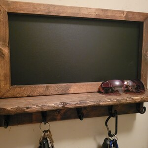May include: A rustic wooden key holder with a chalkboard panel and a shelf. The shelf has three hooks and a pair of sunglasses are resting on the shelf.