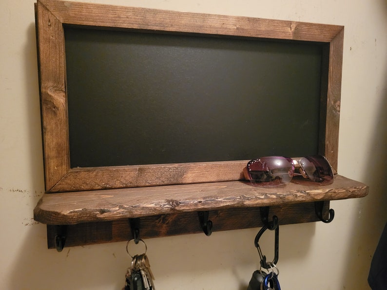 May include: A rustic wooden key holder with a chalkboard panel and a shelf. The shelf has three hooks and a pair of sunglasses are resting on the shelf.