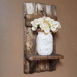 May include: A rustic wooden shelf with a white painted mason jar hanging from a hook. The jar is filled with white flowers and has the text "Ball Mason" on it.