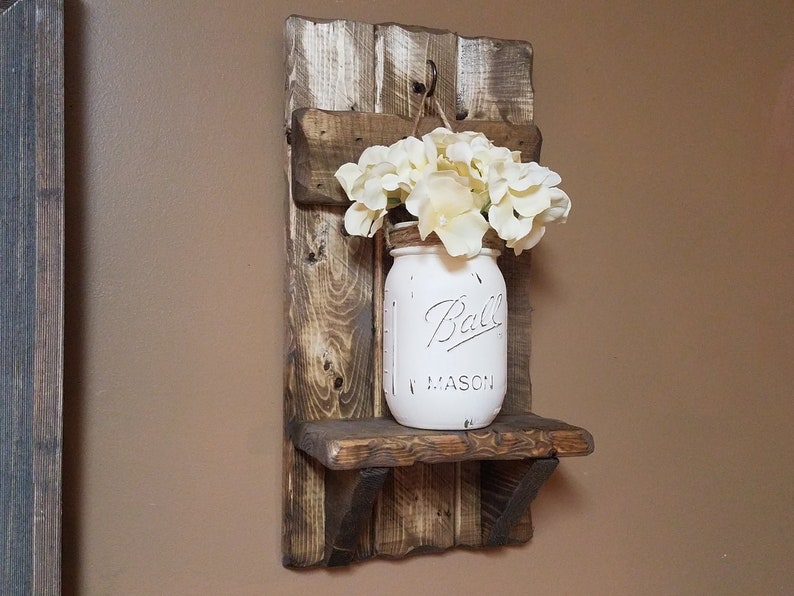 May include: A rustic wooden shelf with a white painted mason jar hanging from a hook. The jar is filled with white flowers and has the text "Ball Mason" on it.