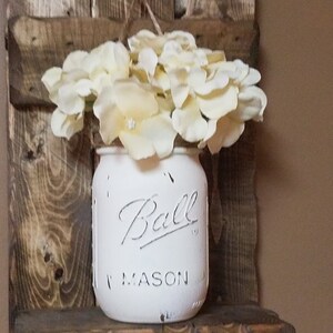 May include: White painted mason jar with the text "Ball Mason" on the front, filled with white flowers, hanging on a rustic wooden shelf.
