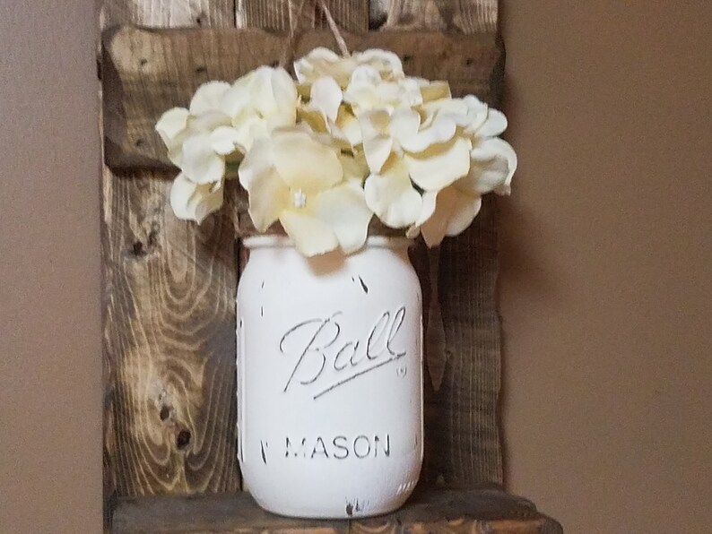 May include: White painted mason jar with the text "Ball Mason" on the front, filled with white flowers, hanging on a rustic wooden shelf.