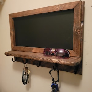 May include: A rustic wooden key holder with a chalkboard panel and hooks. The chalkboard is framed in dark wood and has a shelf below it. There are three hooks on the shelf and a pair of sunglasses are resting on the shelf.