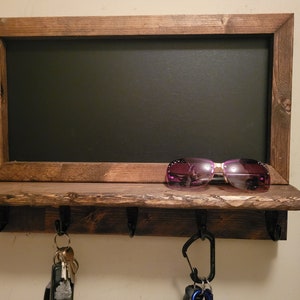 May include: A rustic wooden key rack with a chalkboard above. The rack has four hooks and a shelf. A pair of sunglasses is on the shelf and keys are hanging from the hooks.