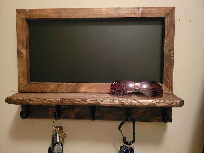 May include: A rustic wooden key rack with a chalkboard above. The rack has four hooks and a shelf. A pair of sunglasses is on the shelf and keys are hanging from the hooks.