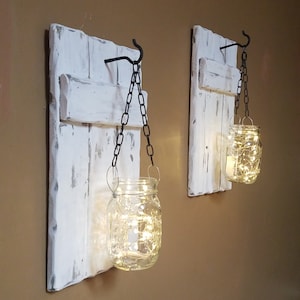 Puede incluir: Dos piezas de decoraci&oacute;n de pared de madera pintadas de blanco con un aspecto r&uacute;stico. Cada pieza tiene un gancho de metal con una cadena que cuelga un frasco de vidrio con luces de hadas en su interior.