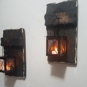 May include: Two black metal lanterns with glass panels and candle holders are mounted on rustic wooden wall plaques. The plaques are stained dark brown and have a white border.